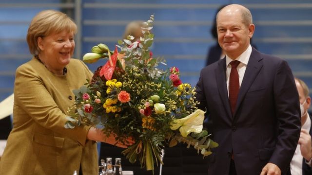 Angela Merkel receiving a bouquet of flowers from Olaf Schulz.