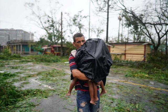 A man carries his son, who is covered in a black raincoat, and everything around him is littered with broken branches and debris.