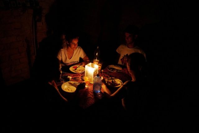 Three members of a family are sitting at the table, everything is dark, they are lit by candles.